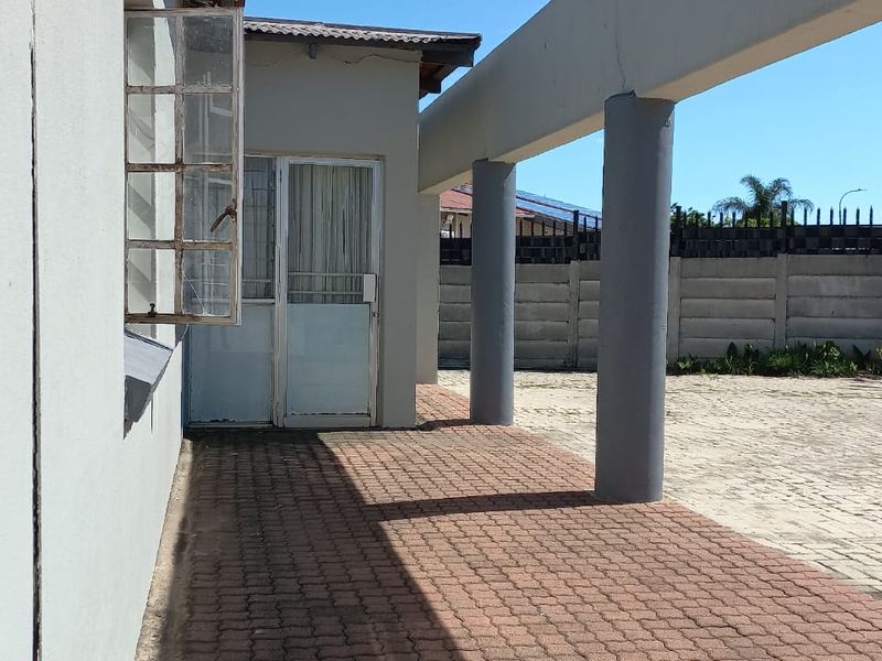 A covered outdoor patio area featuring paved flooring and structural concrete columns, adjacent to a paved driveway and boundary wall.