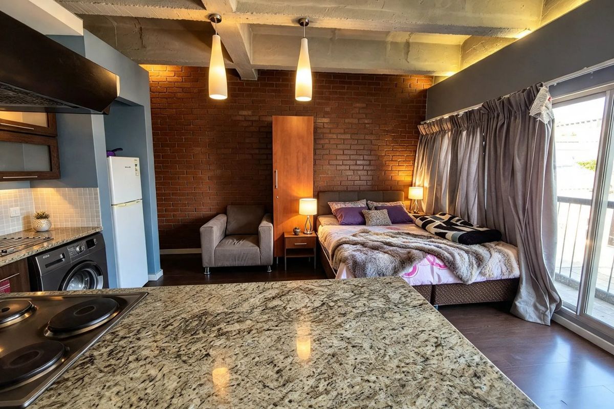 Modern open-plan studio apartment featuring an exposed brick wall, concrete ceiling, granite kitchen countertops, and direct access to a private balcony.