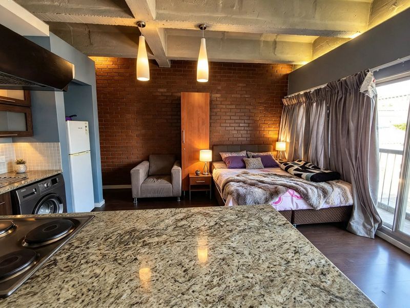 Modern open-plan studio apartment featuring an exposed brick wall, concrete ceiling, granite kitchen countertops, and direct access to a private balcony.