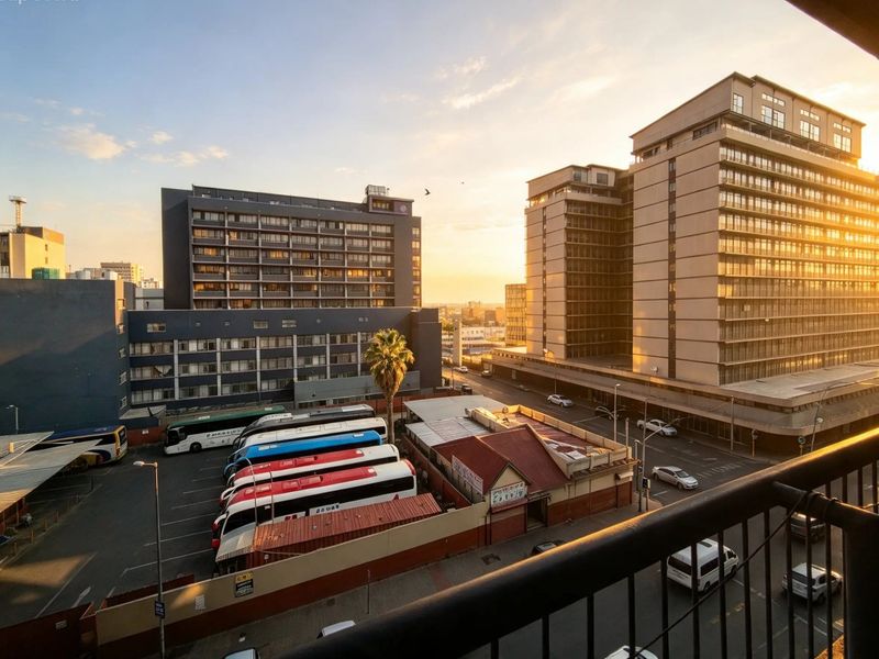 An elevated urban view overlooking a city street, parking area, and surrounding high-rise buildings, captured from a balcony at sunset.