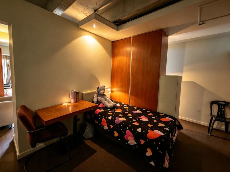 A compact bedroom featuring a built-in wooden wardrobe, exposed concrete ceiling details, and recessed lighting, offering a functional living space.