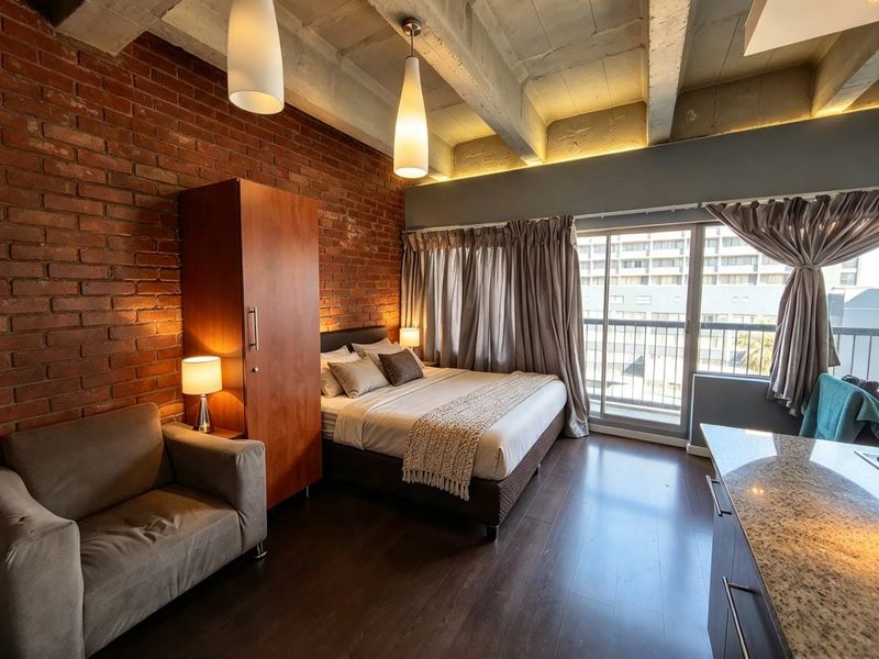 A modern bedroom featuring an exposed brick wall, concrete ceiling beams, and direct access to a private balcony through sliding glass doors.