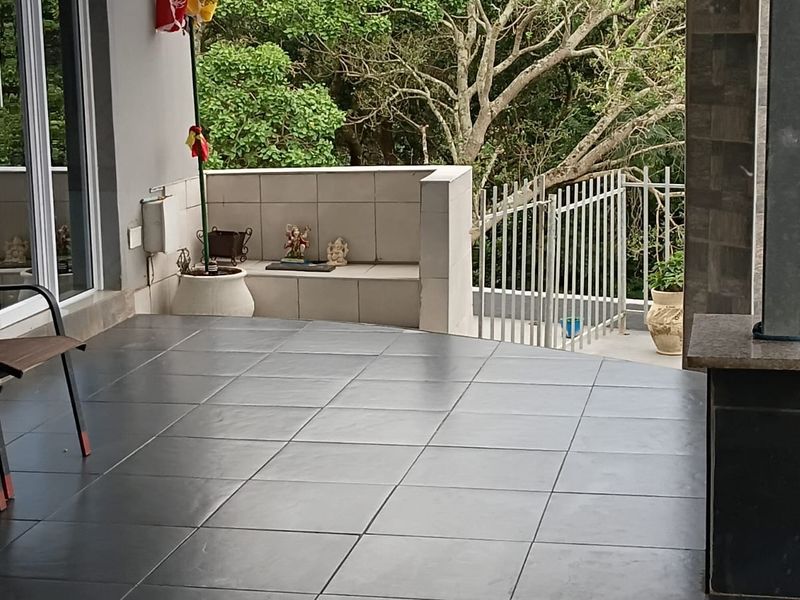 A covered patio area featuring dark tiled flooring, a stone-clad pillar, and a metal balustrade overlooking a lush garden setting.