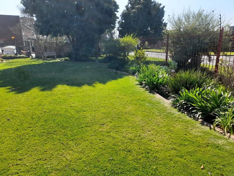 A well-maintained residential garden featuring a lush lawn, established shrubbery, and secure metal perimeter fencing with integrated electric security wires.
