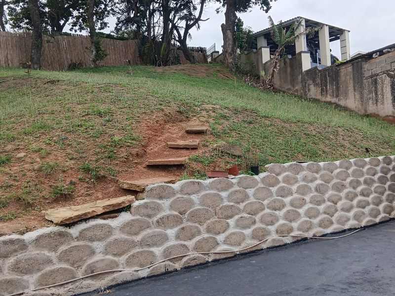 Sloped garden area featuring a decorative concrete retaining wall, stone steps leading up the embankment, and a paved driveway section.