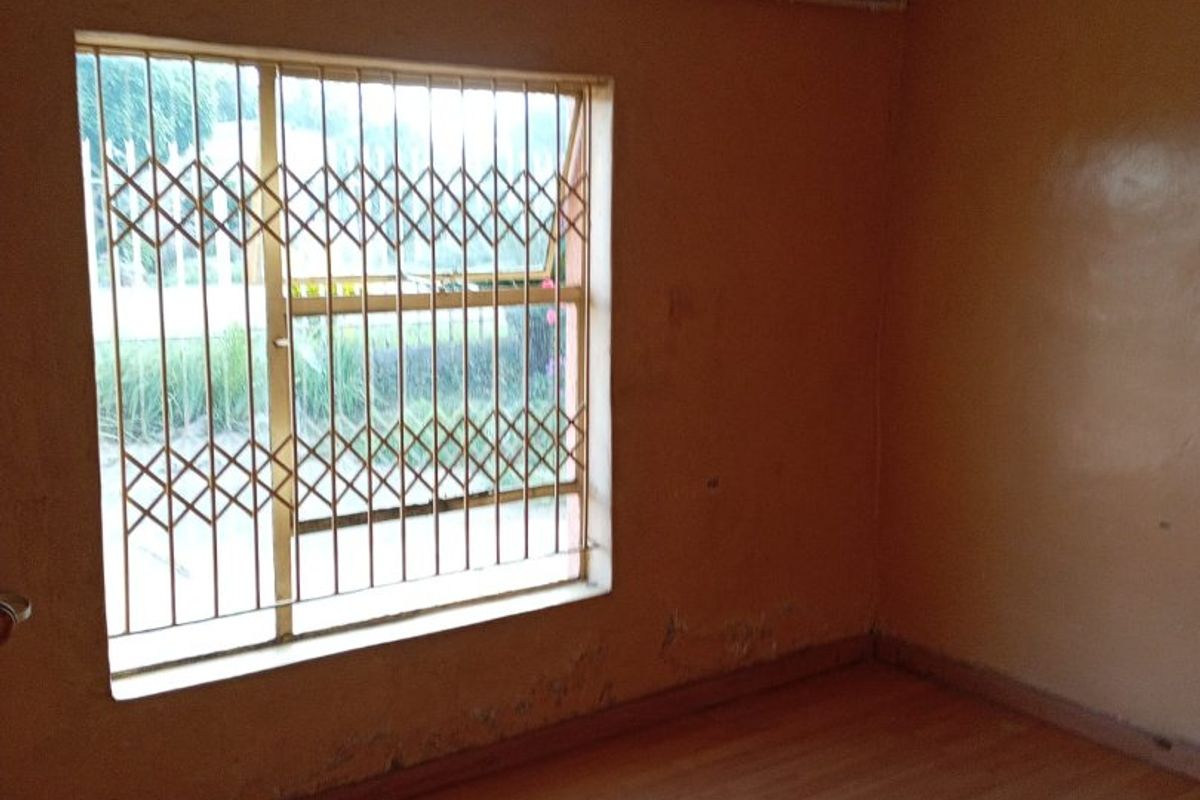 An empty bedroom featuring laminate flooring, a ceiling rose, and a window fitted with a security gate, showing signs of wall surface deterioration.