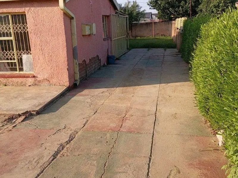Exterior view of a house featuring a concrete driveway, security gates, and visible wear to the exterior wall plaster and driveway surface.