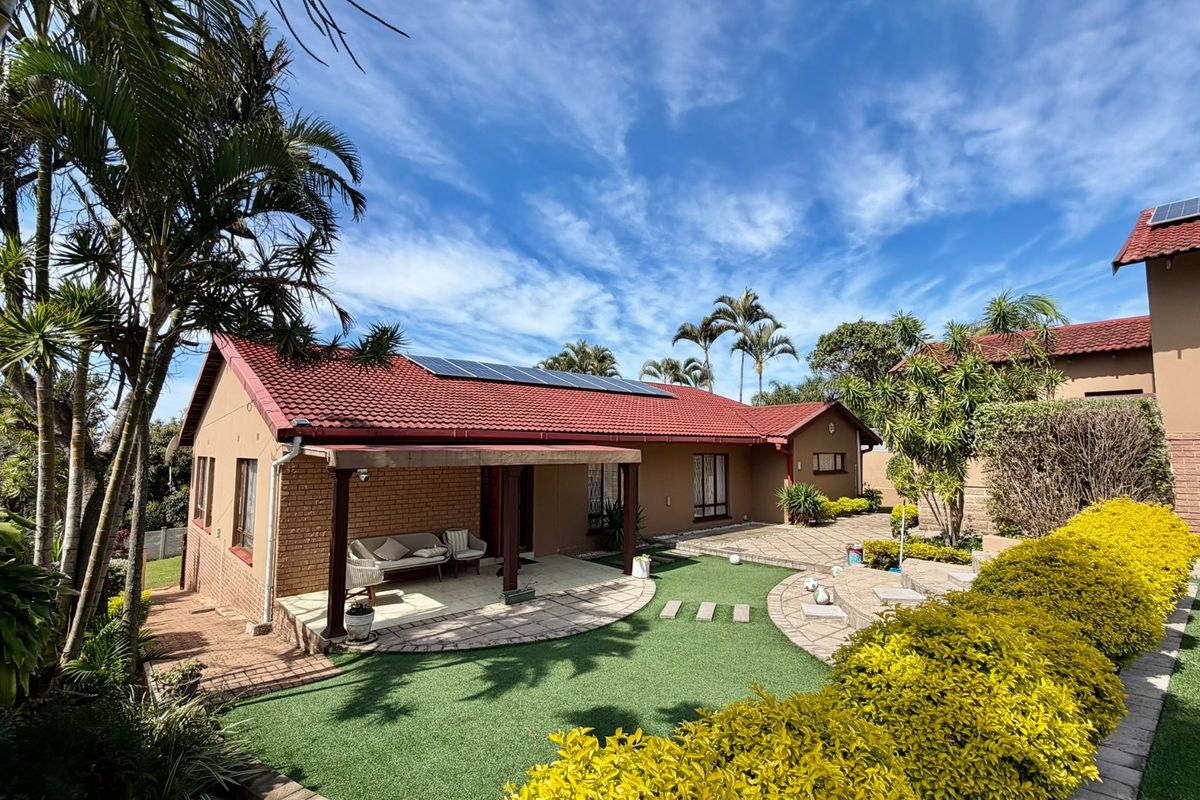 Rear view of the property featuring a covered patio, paved garden areas, artificial lawn, and solar panels installed on the tiled roof.