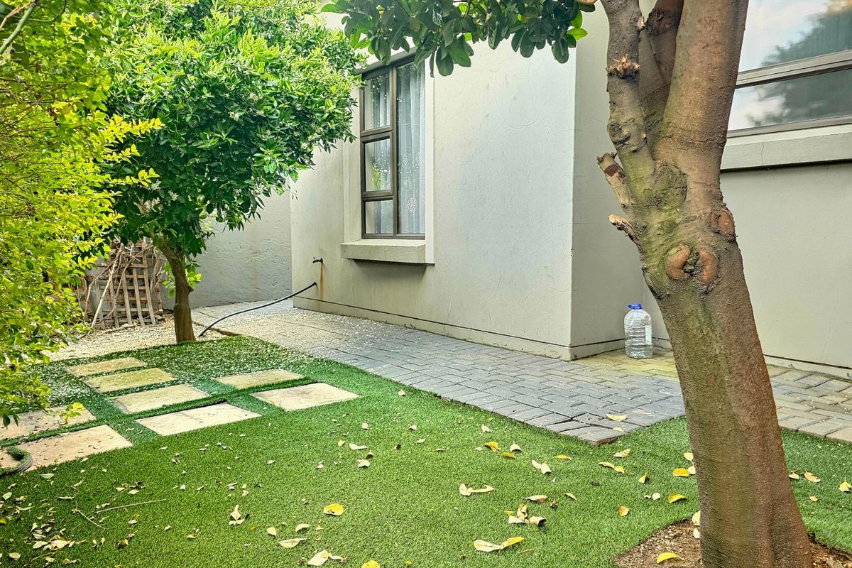 A private rear garden area featuring a paved walkway, artificial lawn, and established trees alongside the exterior wall of the house.