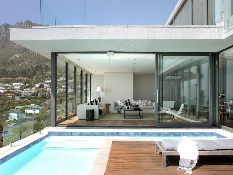 Modern outdoor pool area featuring timber decking, glass balustrades, and seamless indoor-outdoor flow through large sliding glass doors into the living space.