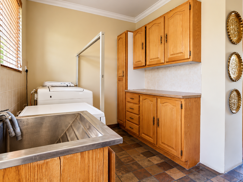 Functional scullery area featuring timber cabinetry, stainless steel sink, tiled splashback, and durable tiled flooring, providing additional workspace and utility storage.