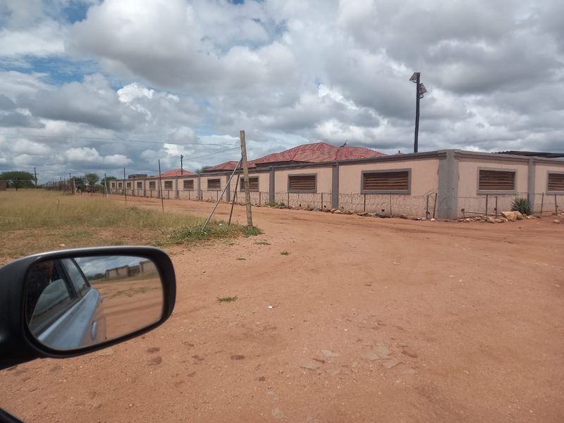 Exterior view of a property featuring a long perimeter wall with security shutters, razor wire fencing, and a tiled roof, set on an unpaved road.