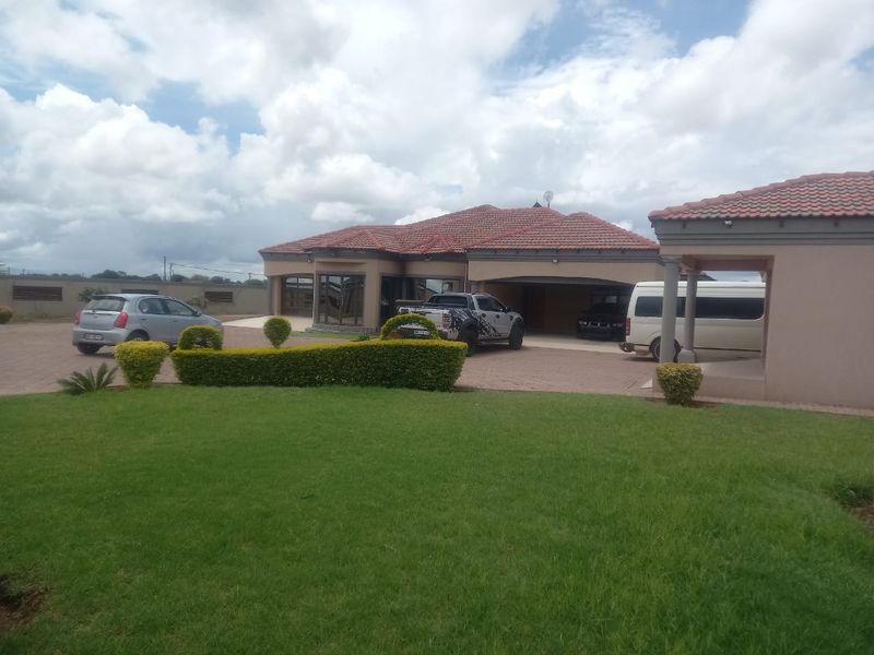 A spacious front exterior view of a residential house featuring a large paved driveway, covered garage, and well-maintained garden area.
