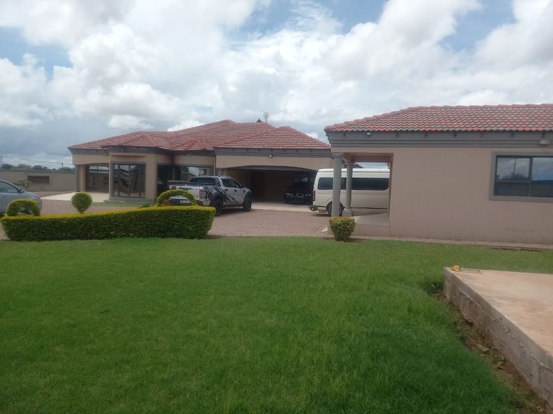 A spacious residential property featuring a large paved driveway, covered patio areas, and well-maintained lawn with manicured hedges.