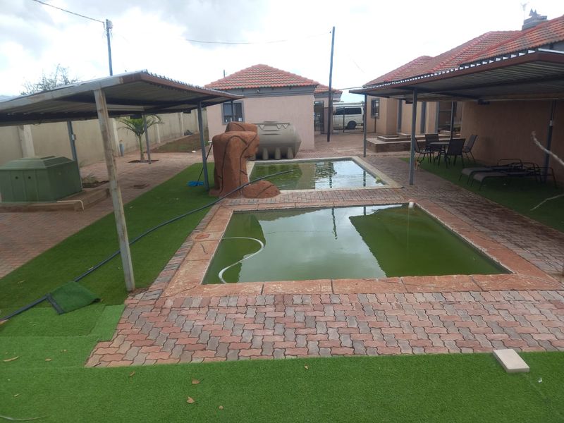 Outdoor pool area featuring two swimming pools, covered patio space, water storage tanks, and paved surrounds with artificial lawn.