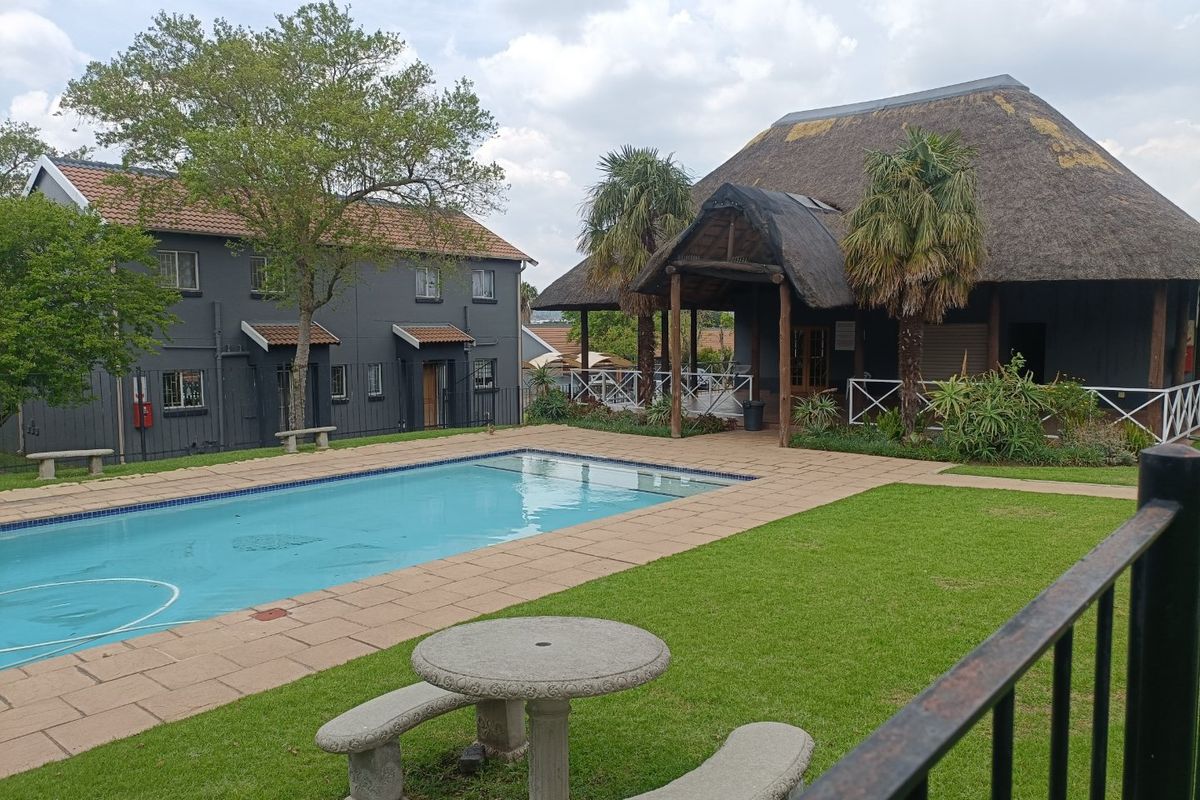 A communal pool area featuring a swimming pool, paved surround, manicured lawn, and a large thatched-roof clubhouse structure.