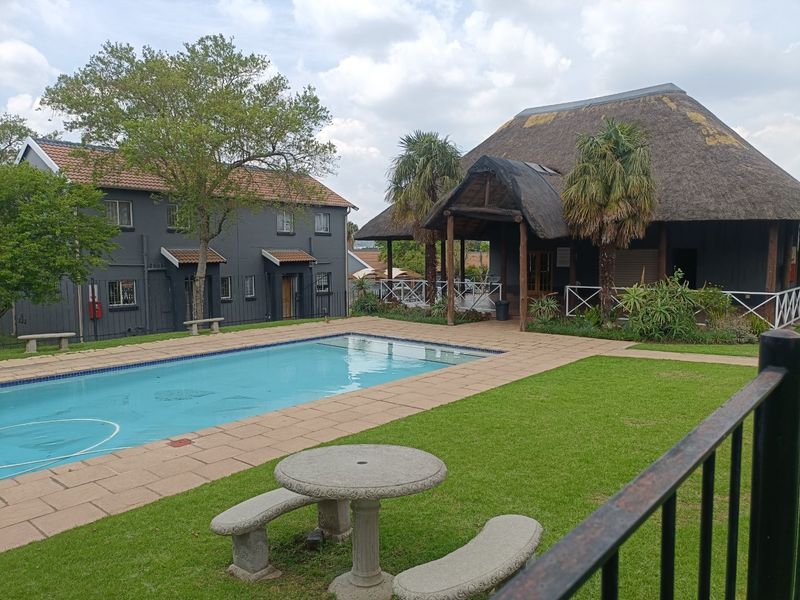 A communal pool area featuring a swimming pool, paved surround, manicured lawn, and a large thatched-roof clubhouse structure.