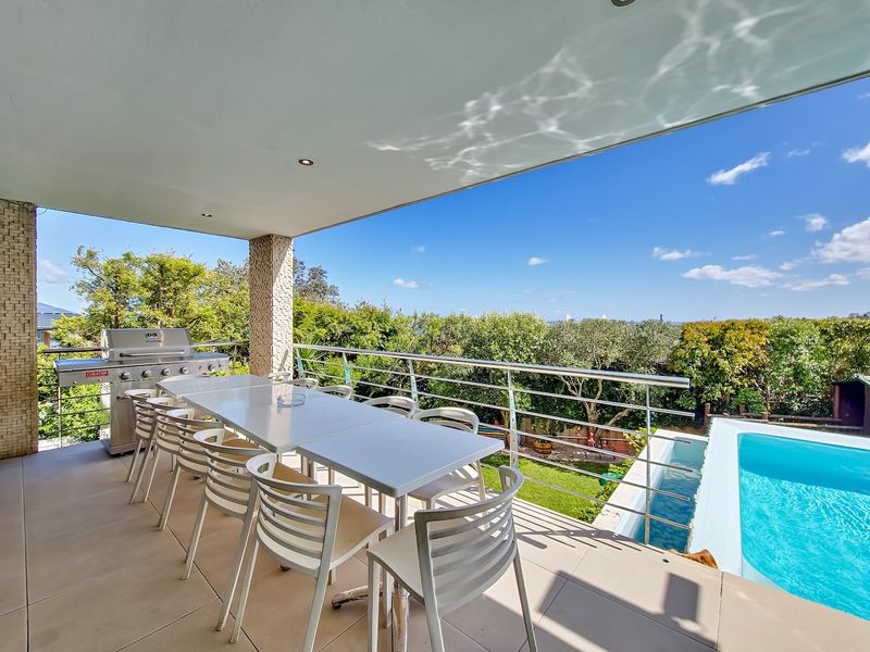 Covered patio featuring tiled flooring, stainless steel balustrade, and built-in gas grill, overlooking a swimming pool and lush garden.