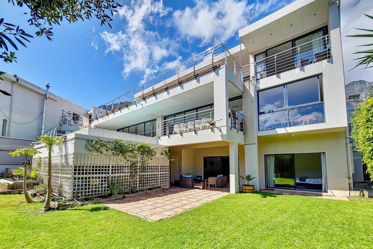 Rear view of a modern multi-story residence featuring expansive balconies with stainless steel railings, a tiled patio area, and well-maintained garden space.