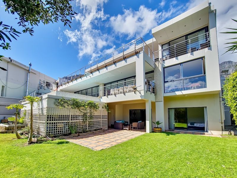 Rear view of a modern multi-story residence featuring expansive balconies with stainless steel railings, a tiled patio area, and well-maintained garden space.