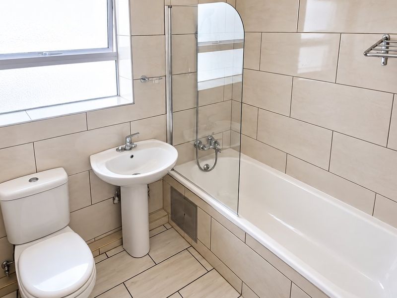 A clean, modern bathroom featuring a pedestal basin, toilet, and a bathtub with a glass shower screen, finished with neutral wall and floor tiles.