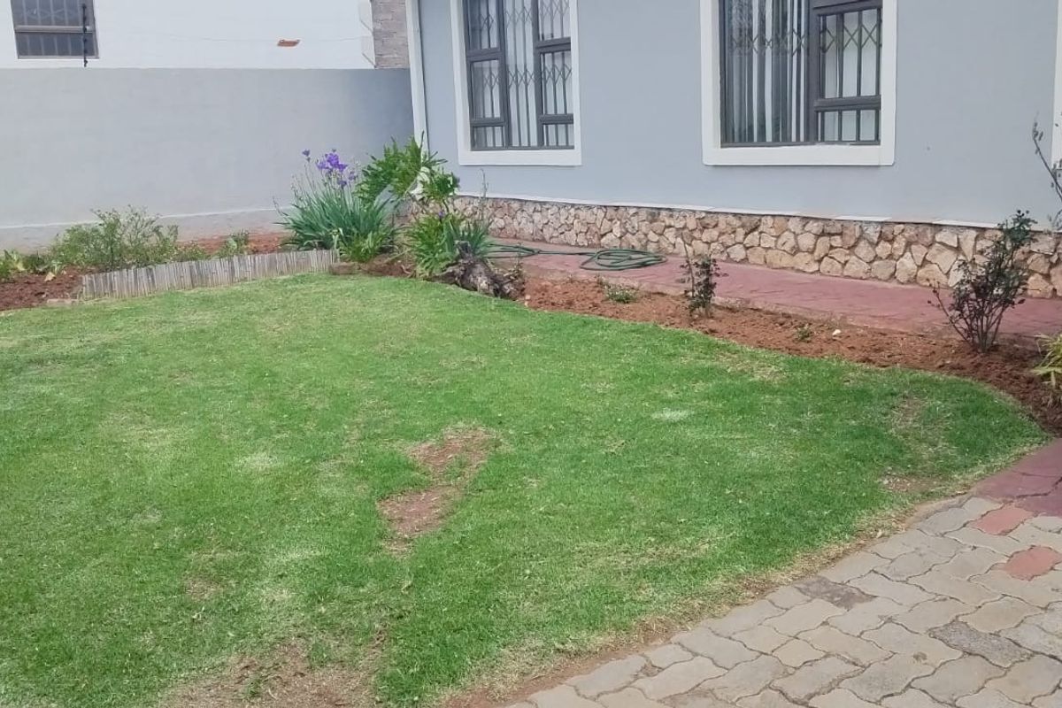 A rear garden area featuring a lawn, stone-clad exterior wall, security bars on windows, and paved access path.