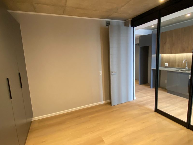 Modern open-plan apartment featuring an exposed concrete ceiling, light wood laminate flooring, built-in storage, and a sleek kitchen separated by a sliding glass partition.