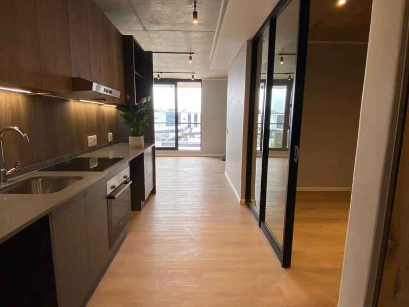 Modern open-plan kitchen featuring sleek cabinetry, integrated appliances, and exposed concrete ceilings, leading to a bright living area with sliding glass doors.