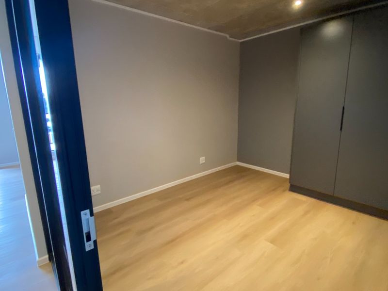 A modern, empty bedroom featuring light laminate flooring, an exposed concrete ceiling, and a dark grey built-in wardrobe.