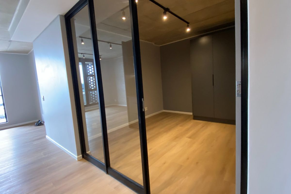 A modern, empty bedroom featuring sleek sliding glass doors, built-in wardrobes, track lighting, and an exposed concrete ceiling with light wood-look flooring.