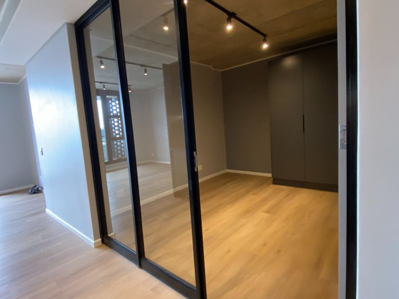A modern, empty bedroom featuring sleek sliding glass doors, built-in wardrobes, track lighting, and an exposed concrete ceiling with light wood-look flooring.