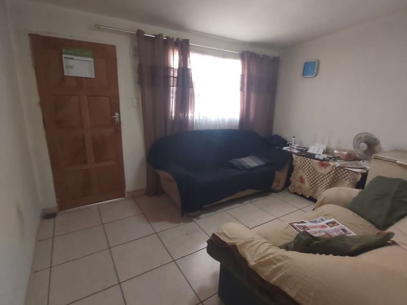 A living room featuring tiled flooring, a wooden entrance door, and a window with curtains, providing a basic functional living space.