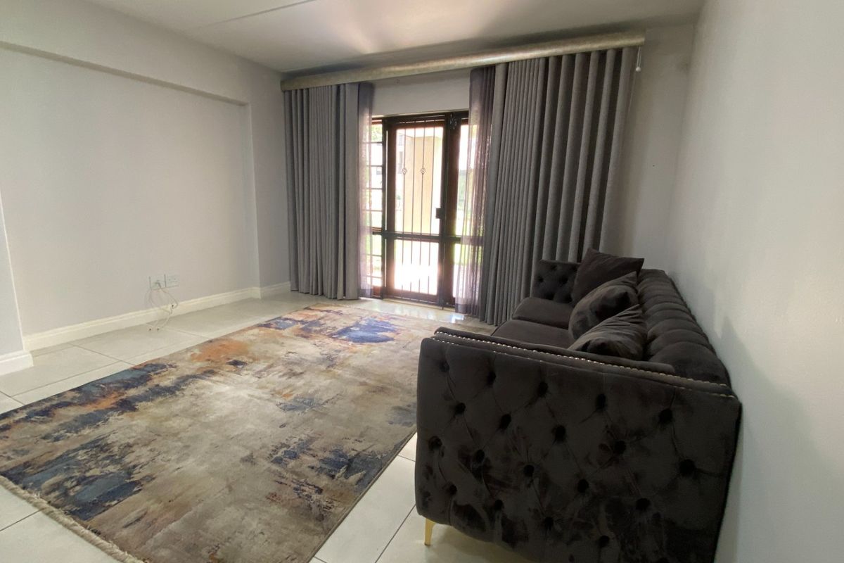 A bright living room featuring neutral tiled flooring, large windows with curtain tracks, and a secure metal entrance gate leading to the exterior.