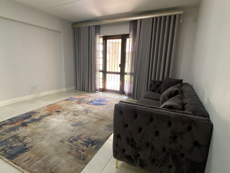 A bright living room featuring neutral tiled flooring, large windows with curtain tracks, and a secure metal entrance gate leading to the exterior.