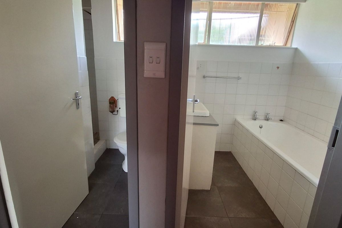 A functional bathroom featuring a built-in bathtub, vanity basin, and separate toilet area with neutral wall and floor tiling.