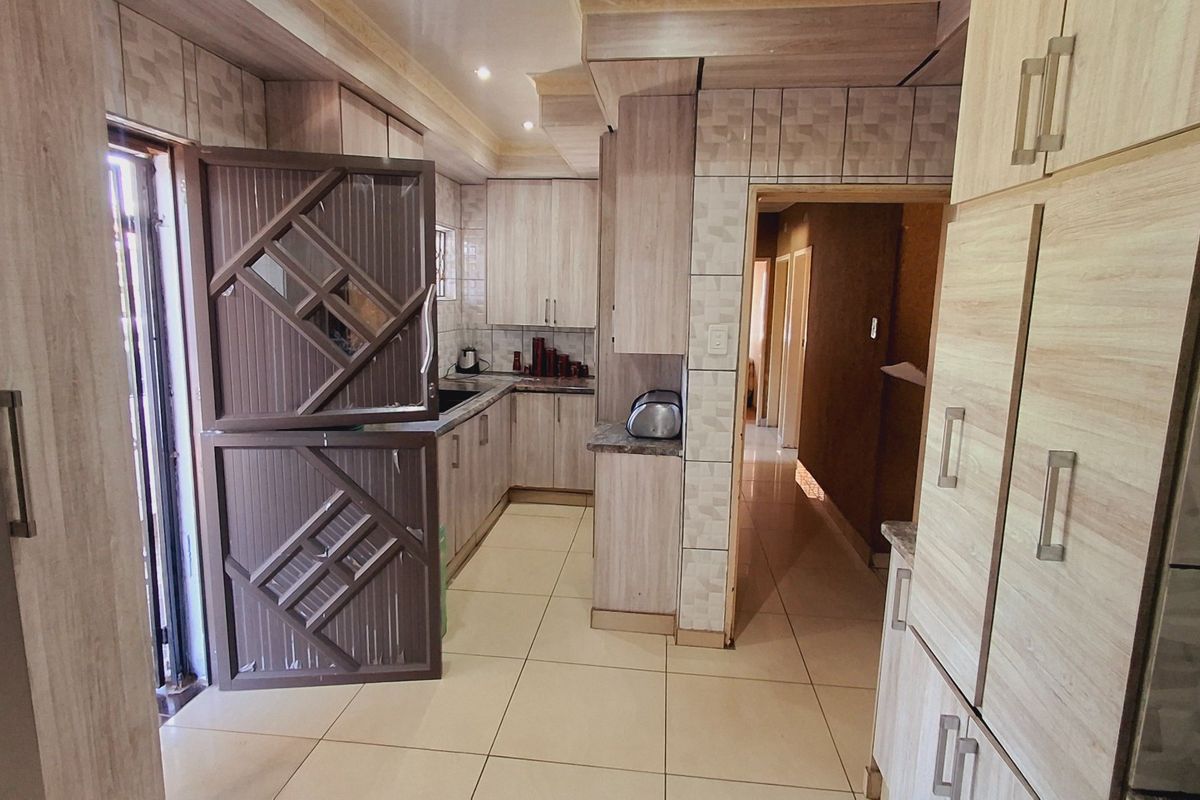 A functional kitchen featuring light wood-finish cabinetry, tiled flooring, and a tiled backsplash, with direct access to an adjacent hallway.
