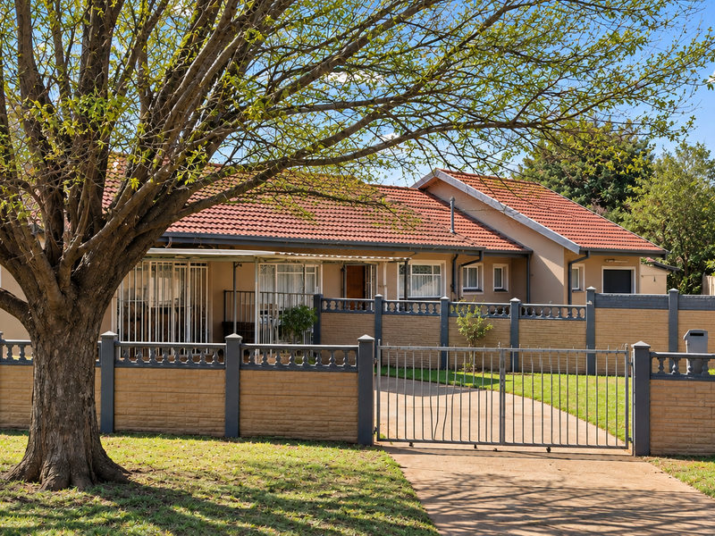 Front view of a single-story house featuring a tiled roof, paved driveway, and a secure perimeter wall with a metal gate.