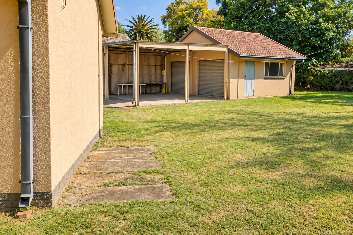 Rear view of the property featuring a spacious lawn, a detached garage with a carport, and established garden surroundings.