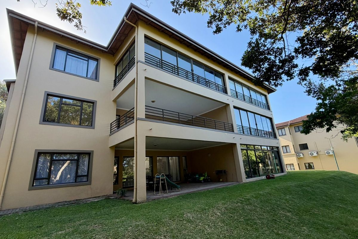 Rear view of a multi-story apartment building featuring stacked balconies, large glass windows, and a ground-level paved patio area overlooking a lawn.