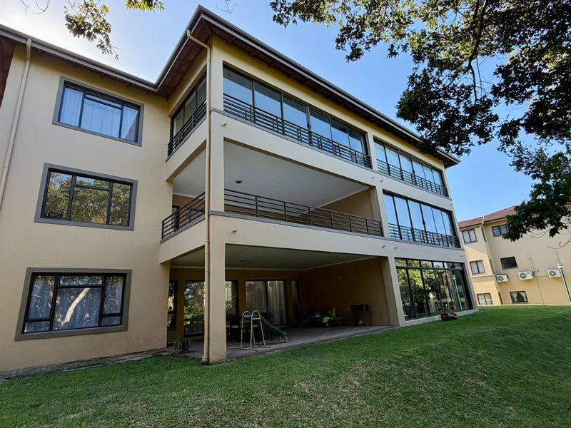 Rear view of a multi-story apartment building featuring stacked balconies, large glass windows, and a ground-level paved patio area overlooking a lawn.