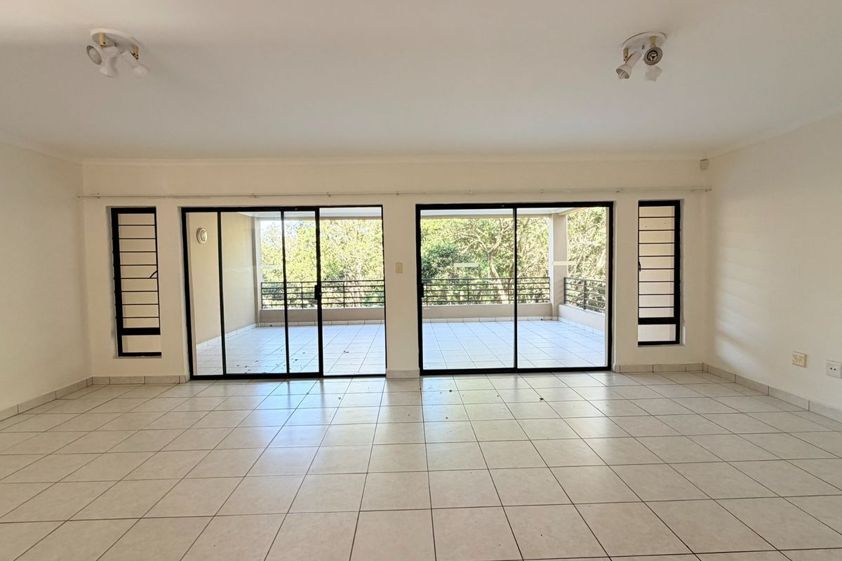 Spacious, empty living area featuring neutral tiled flooring and large sliding glass doors that open onto a private balcony with natural views.