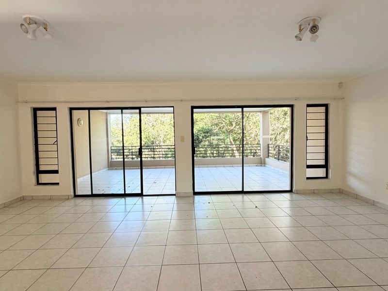 Spacious, empty living area featuring neutral tiled flooring and large sliding glass doors that open onto a private balcony with natural views.