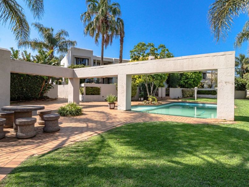 Outdoor pool area featuring a paved patio, concrete pergola structure, and a well-maintained garden setting.