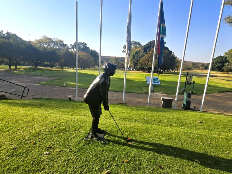 A well-maintained golf course area featuring a decorative golfer statue, multiple flagpoles, and paved pathways set within a landscaped green environment.