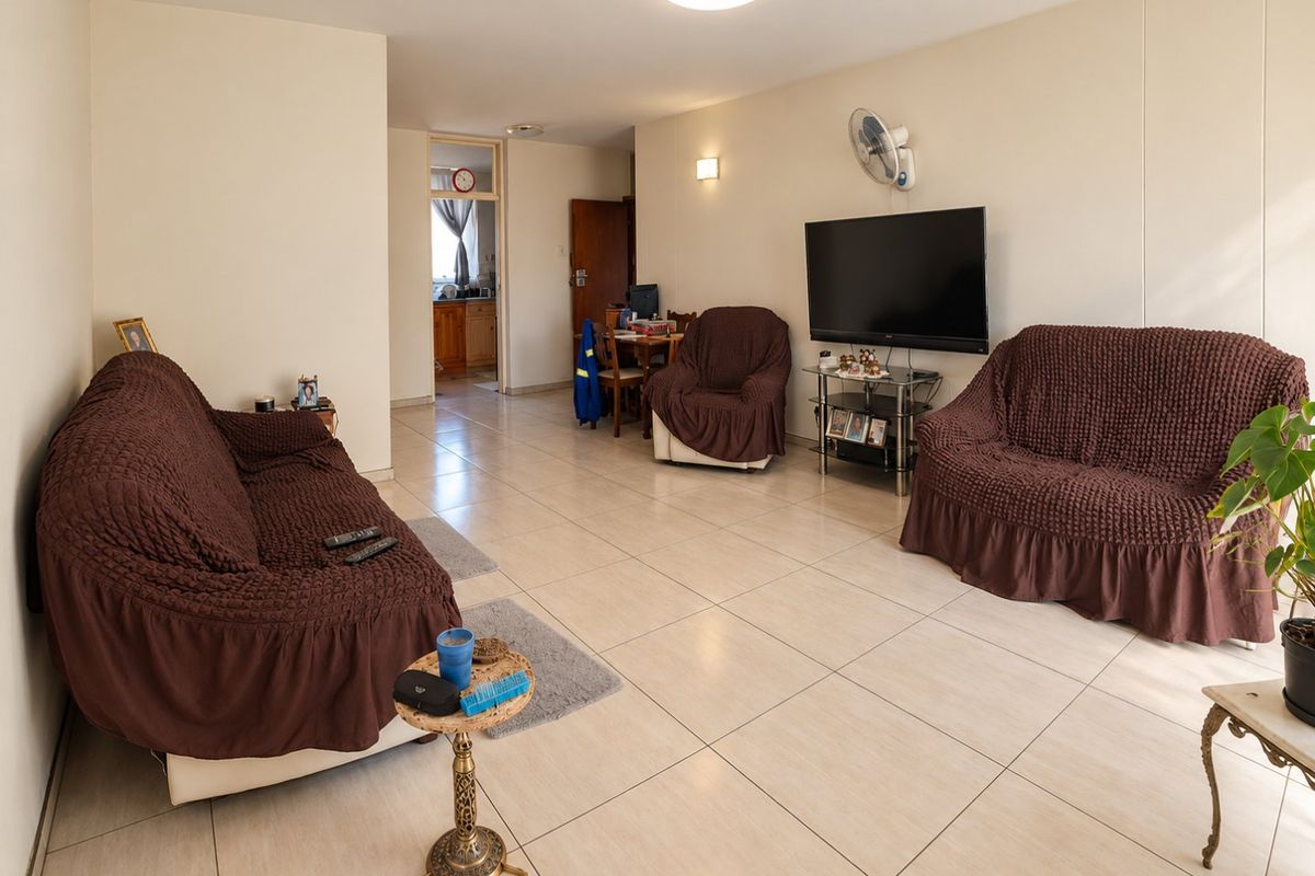 A spacious living room featuring neutral tiled flooring, a wall-mounted fan, and direct access to the adjacent kitchen area.