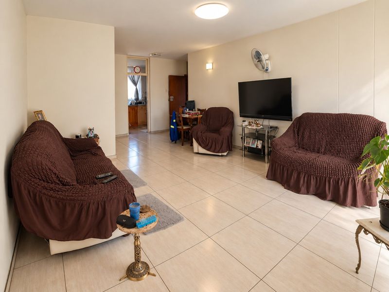 A spacious living room featuring neutral tiled flooring, a wall-mounted fan, and direct access to the adjacent kitchen area.