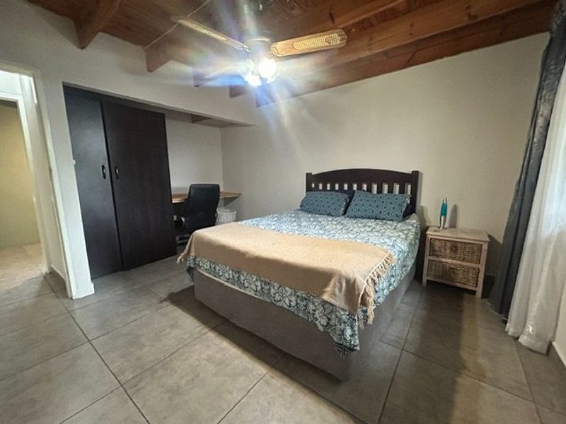 A bedroom featuring tiled flooring, exposed timber ceiling beams, a ceiling fan, and a built-in wardrobe with an integrated desk area.
