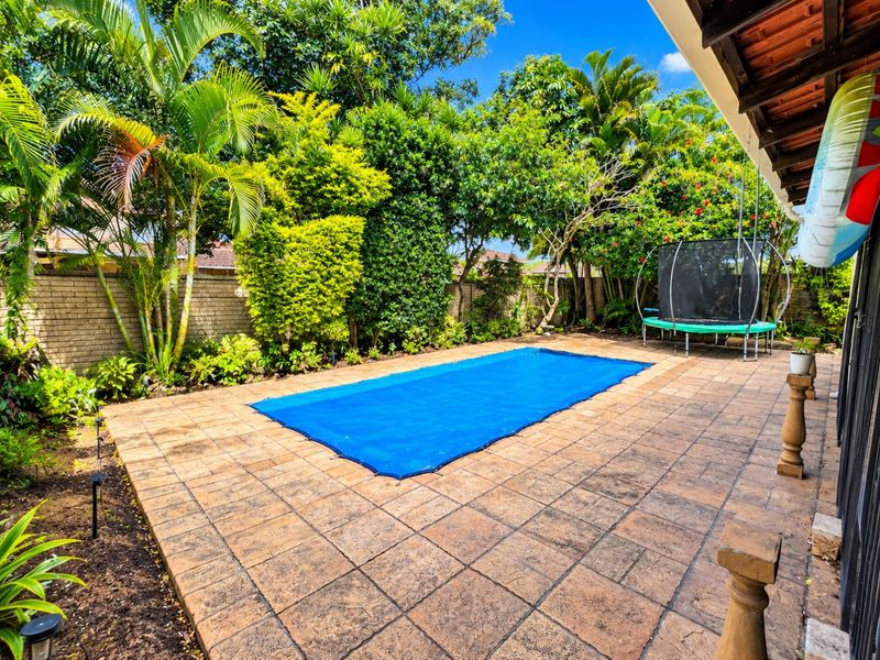 A well-maintained outdoor pool area featuring a paved patio, a blue pool cover, and lush garden landscaping with a brick boundary wall.