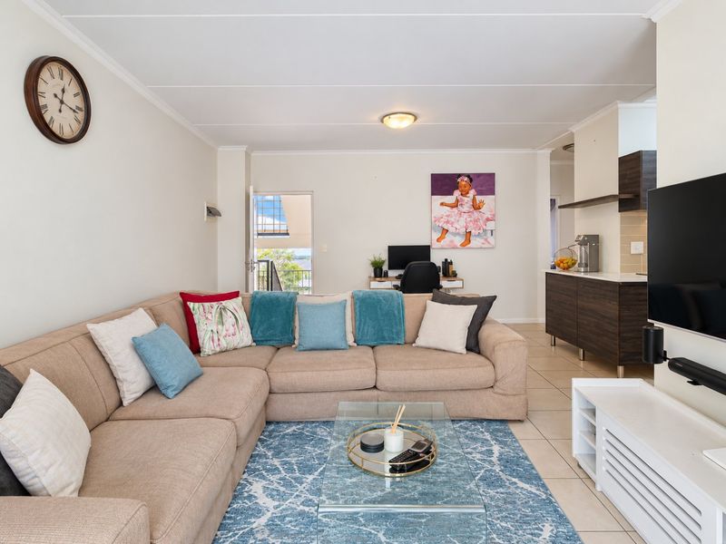 An open-plan living area featuring tiled floors, neutral walls, and a modern kitchen corner with dark cabinetry and stone countertops, leading to a balcony.