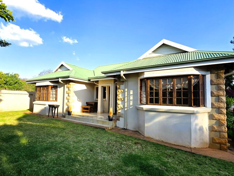 Rear view of a charming house featuring a green corrugated metal roof, stone-clad accents, timber bay windows, and a paved patio overlooking the garden.